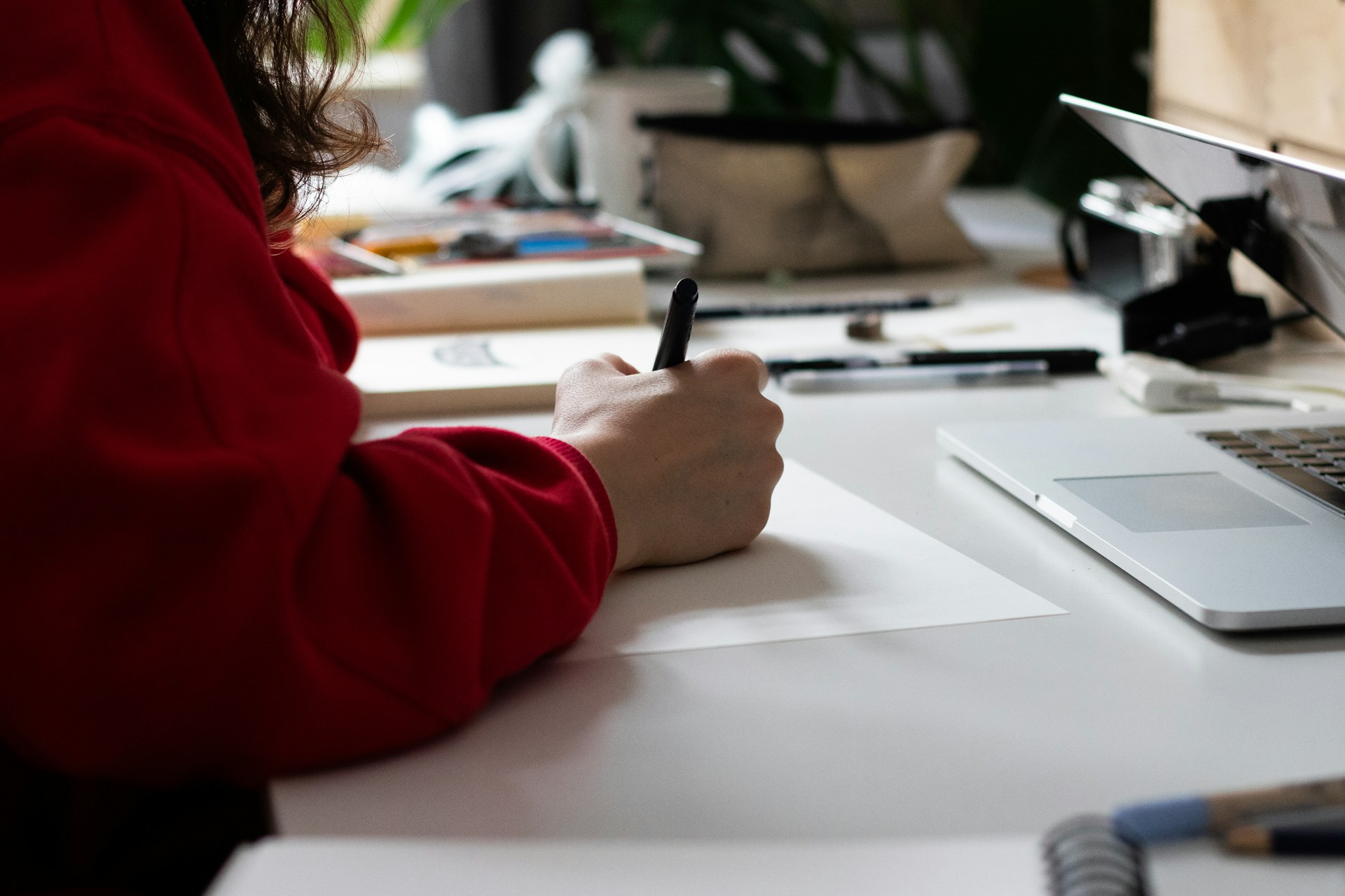 Close-up of a person in a red sweater writing with a black pen on white paper at a clean desk, with a laptop nearby.